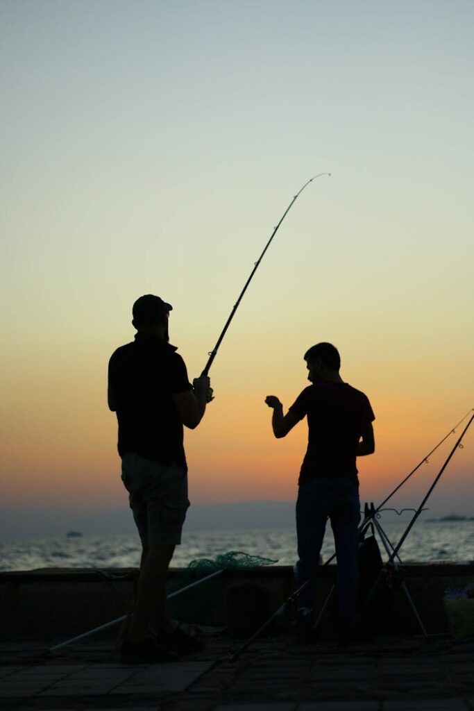Two anglers silhouetted against a dramatic sunset as they fish by the sea.